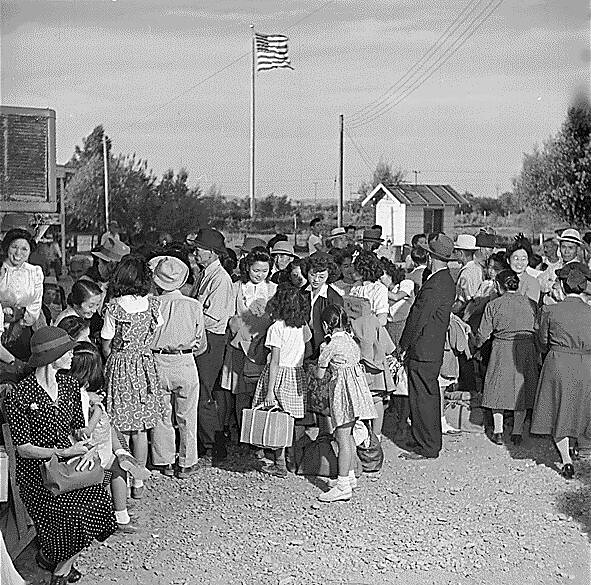 People are gathered at a bus stop some stand, some sit. Photo is black and white.