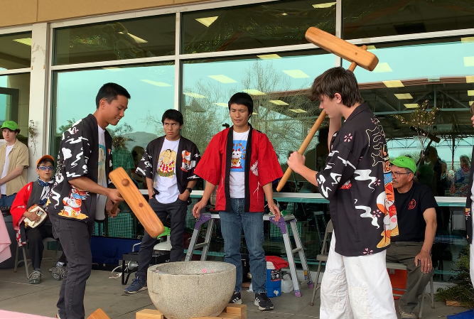 Two young men in traditional Japanese costume, swing large wooden mochi hammers down into a heavy large bowl.