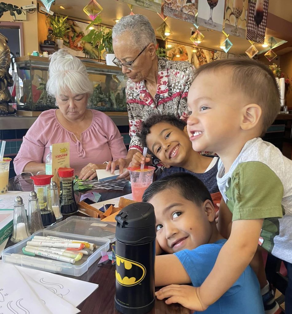 Two elderly women and 3 young boys work on  origami together